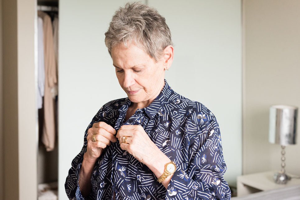 Waist up view of older woman doing up buttons on blue blouse while getting dressed in bedroom (selective focus)
