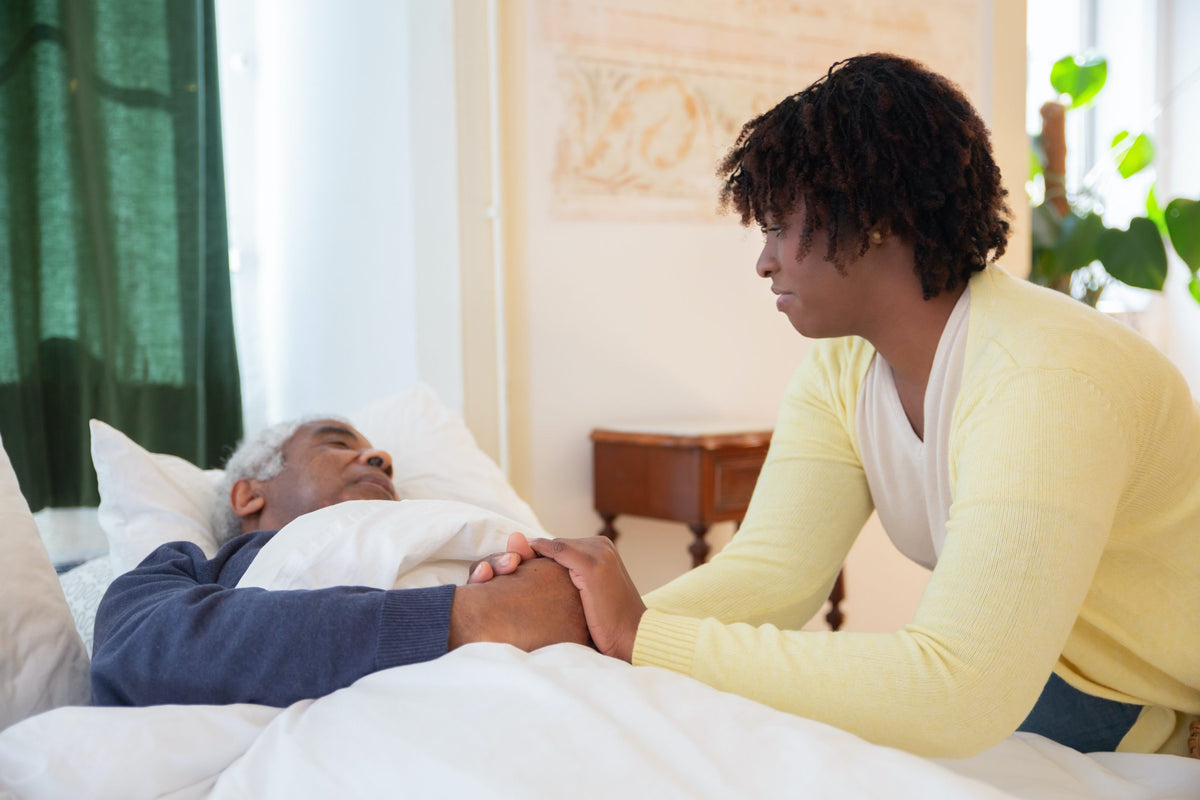 Young women sitting beside bed and holding hand of older man laying down.