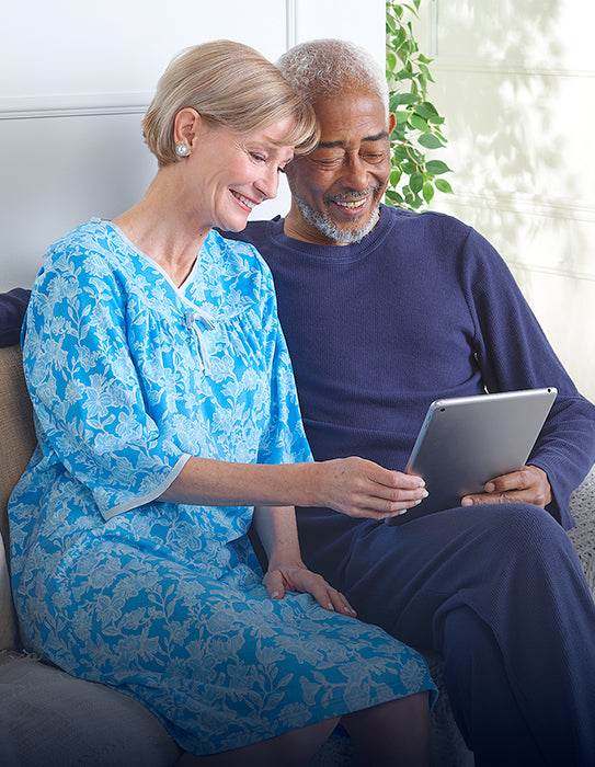 Smiling older couple seated together on a sofa, looking at a tablet while wearing comfortable adaptive clothing.