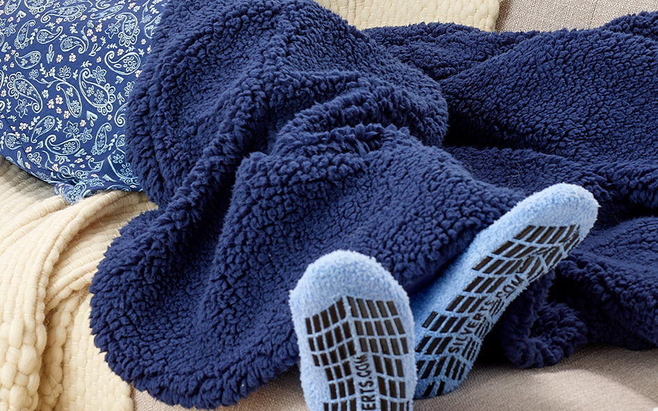 Person wrapped in a navy fleece blanket with light blue non-slip grip socks resting on a sofa, creating a warm and cozy setting.
