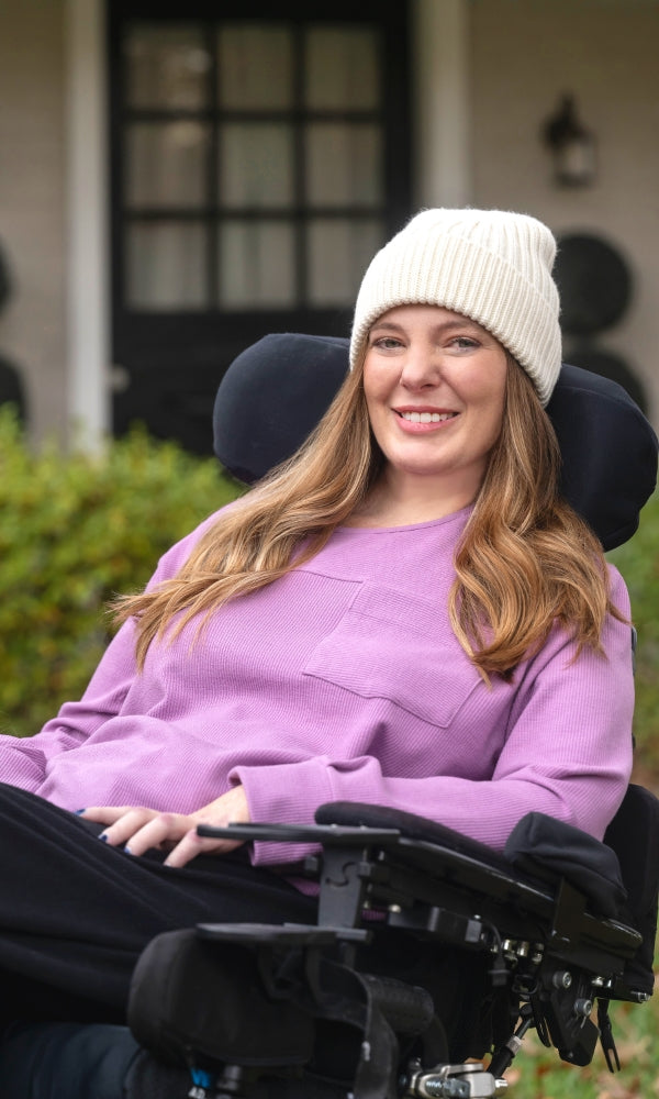 Woman in a wheelchair wearing a purple top and white knit hat outdoors.