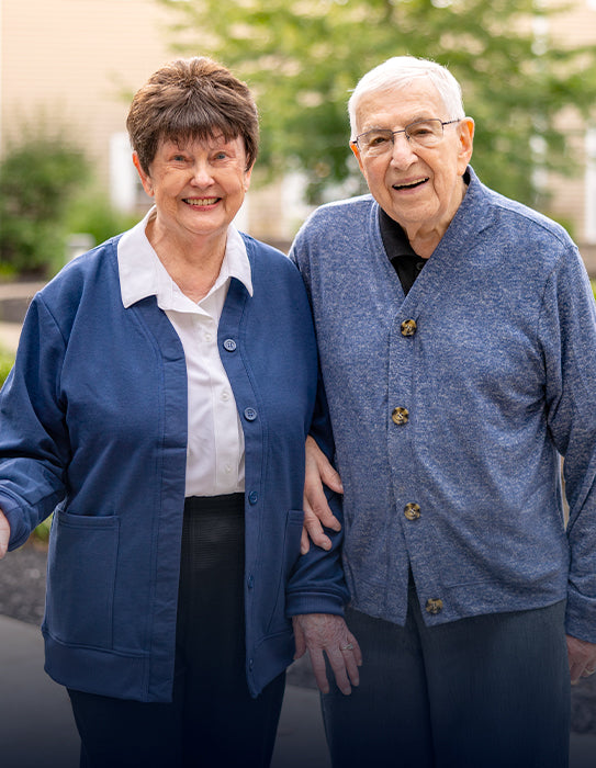 Smiling older couple standing outdoors wearing adaptive clothing, including a blue adaptive cardigan and a blue adaptive button-front sweater.